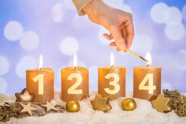 Four numbered gold advent candles with four candles lit by hand with match and christmas decoration lying in snow as template