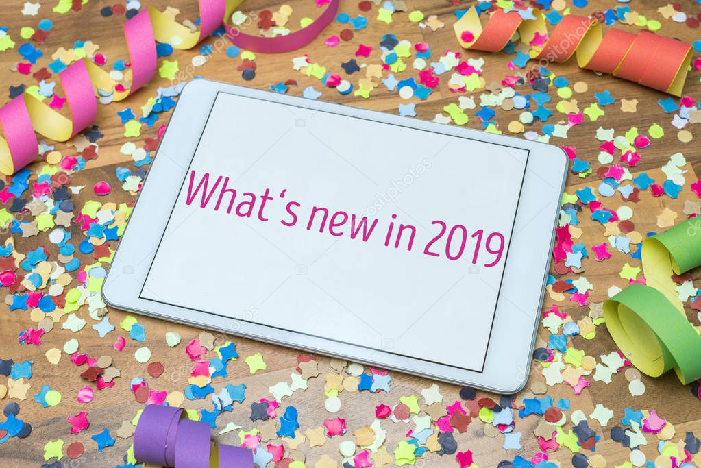 Colorful confetti and paper streamers on wooden table in background with white tablet and What's new in 2019 message written on display