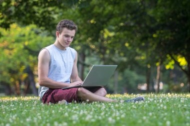 Parktaki genç adam çimlerin üzerinde dizüstü bilgisayarla oturuyor..