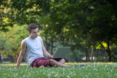 Parktaki genç adam çimlerin üzerinde dizüstü bilgisayarla oturuyor..