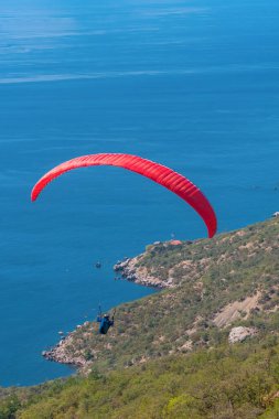 Bir adam, açık bir yaz gününde yeşil bir dağ ve mavi bir deniz üzerinde bir paraglider üzerinde uçar.