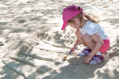 Küçük bir kız arkeoloğu oynuyor, açık bir günde dinazor iskeletinin modelini kazıyor.