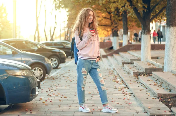 Mulher bonita em jeans e uma camisola com uma mochila com café — Fotografia de Stock