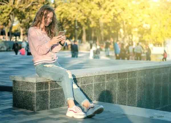 Mulher sentada jeans sweatshirt tênis assistir smartphone fone de ouvido do telefone móvel — Fotografia de Stock