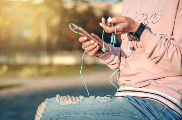 Mulher sentada jeans sweatshirt assistir smartphone fones de ouvido do telefone celular na rua da cidade — Fotografia de Stock
