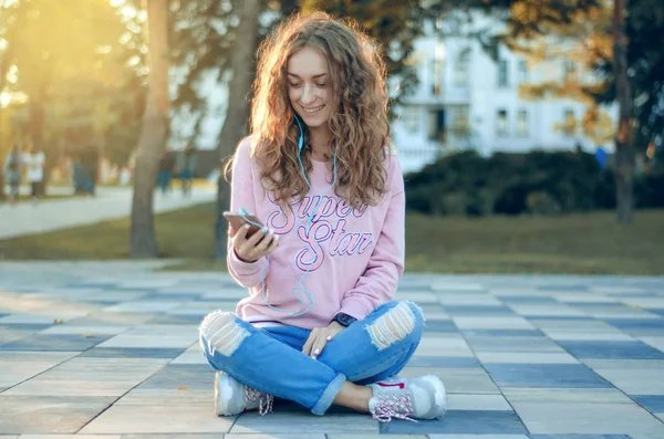 Mulher sentada jeans sweatshirt tênis assistir smartphone fone de ouvido celular selfie feliz sorrindo — Fotografia de Stock