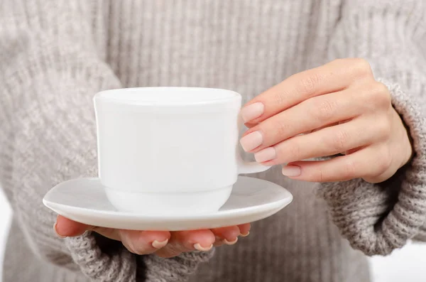 Jeune femme en pull tenant une tasse blanche et une soucoupe dans les mains, buvant du café chaud . — Photo