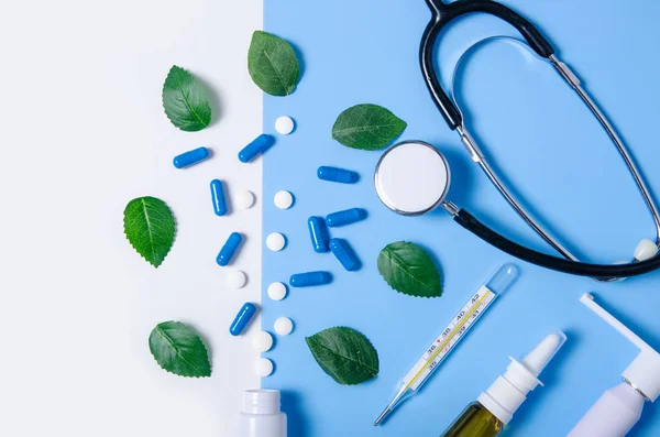 Composition nasal spray, throat spray, stethoscope, white jar with pills, thermometer nature green leaf on color blue white background, flat lay, top view