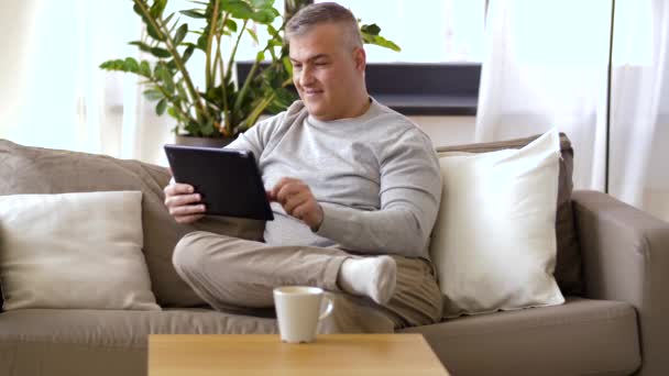 Muž s tablet pc sedí na gauči doma — Stock video