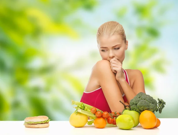 Zweifelnde Frau mit Früchten schaut auf Hamburger — Stockfoto