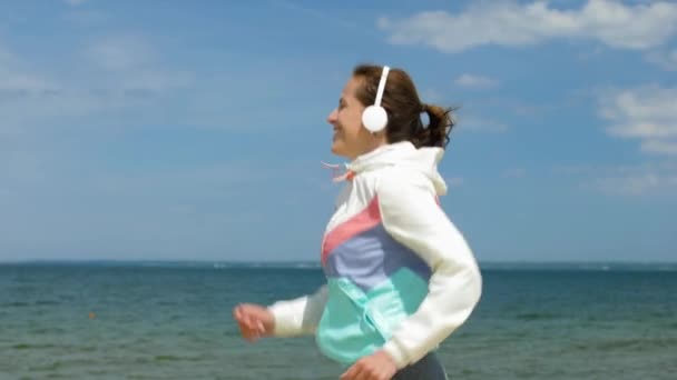 Usmívající se žena se sluchátky běží podél pláže — Stock video
