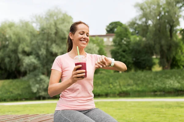 Žena s koktejl při pohledu na chytré hodinky v parku — Stock fotografie