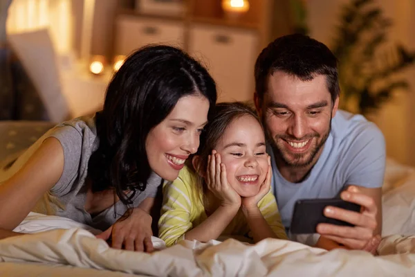 Familia feliz con teléfono inteligente en la cama por la noche —  Fotos de Stock