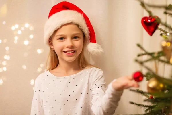 Happy girl in santa hat decorating christmas tree — Stock Photo, Image