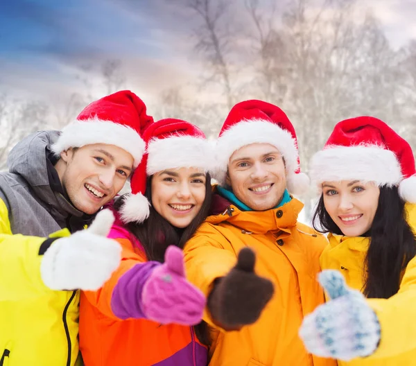 Přátelé v santa klobouky ukazuje palec nahoru venku — Stock fotografie