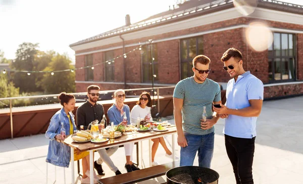 休闲和人的概念 快乐的朋友与智能手机在屋顶烧烤派对 — 图库照片