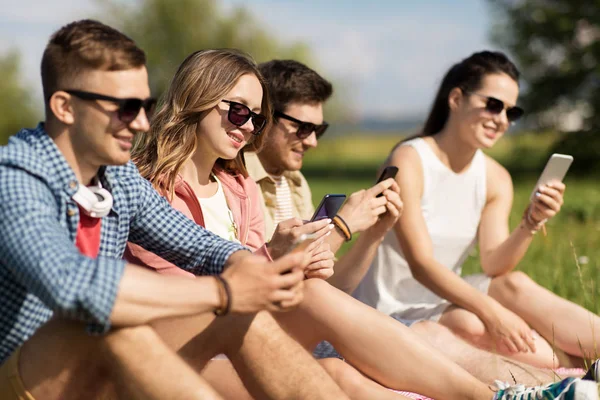 Amigos sonrientes con teléfonos inteligentes sentados en la hierba — Foto de Stock