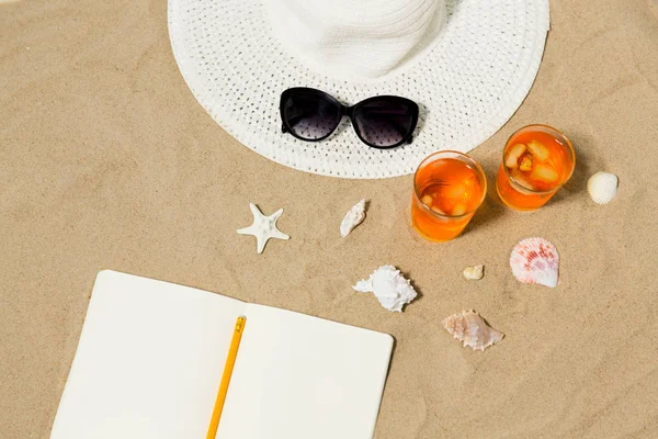Caderno, coquetéis, chapéu e sombras na areia da praia — Fotografia de Stock