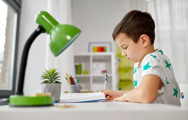 Student pojke med bok skriva till anteckningsbok hemma — Stockfoto