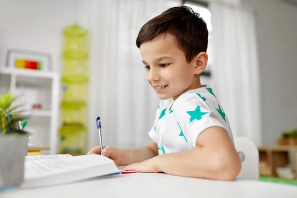 Estudante menino com livro escrevendo para notebook em casa — Fotografia de Stock