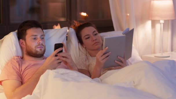 Casal com smartphone e livro na cama à noite — Vídeo de Stock