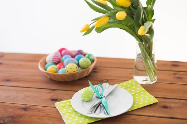 Huevos de Pascua en cesta, platos, cubiertos y flores — Foto de Stock