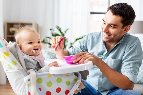 Отец кормит счастливого ребенка в высокий детский стульчик на дому — стоковое фото