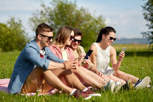 Улыбающиеся друзья со смартфонами, сидящими на траве — стоковое фото