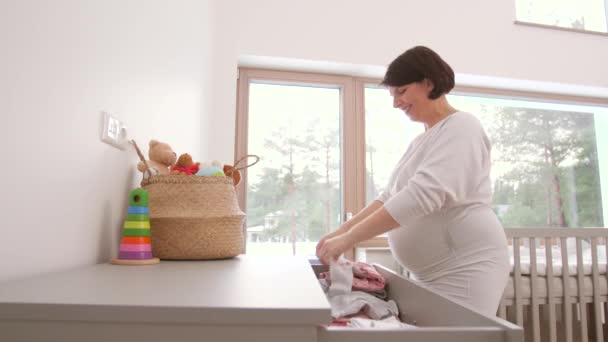 Heureuse femme enceinte réglage vêtements de bébé à la maison — Video