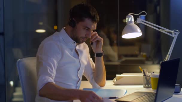 Hombre de negocios en auriculares con ordenador portátil en la oficina de noche — Vídeo de stock