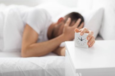 close up of man in bed reaching for alarm clock clipart