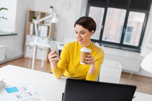 Счастливая женщина с кофе с помощью смартфона в офисе — стоковое фото