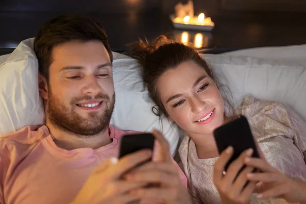 Casal feliz usando smartphones na cama à noite — Fotografia de Stock