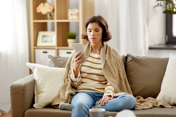 Грустная больная женщина в одеяле с помощью смартфона дома — стоковое фото