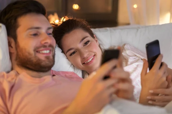 Casal feliz usando smartphones na cama à noite — Fotografia de Stock