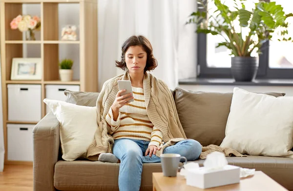 Battaniyeli üzgün hasta kadın evde akıllı telefon kullanıyor. — Stok fotoğraf
