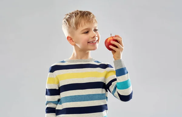 Улыбающийся мальчик в полосатом свитере ест красное яблоко — стоковое фото