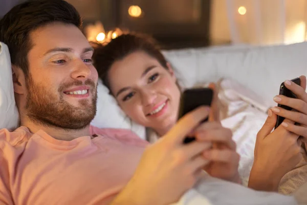 Pareja feliz usando teléfonos inteligentes en la cama por la noche — Foto de Stock
