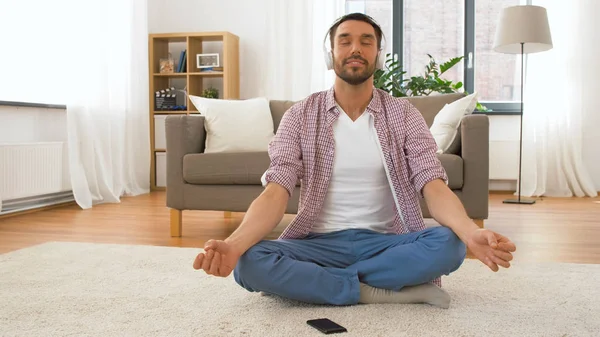 Homem em fones de ouvido meditando em lótus posar em casa — Fotografia de Stock