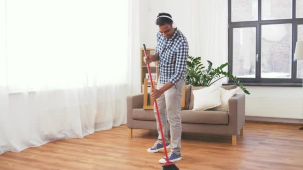 Man in hoofdtelefoon met bezem schoonmaken thuis — Stockvideo