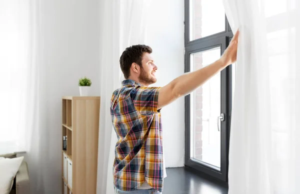 Jeune homme ouvrant rideau de fenêtre à la maison — Photo