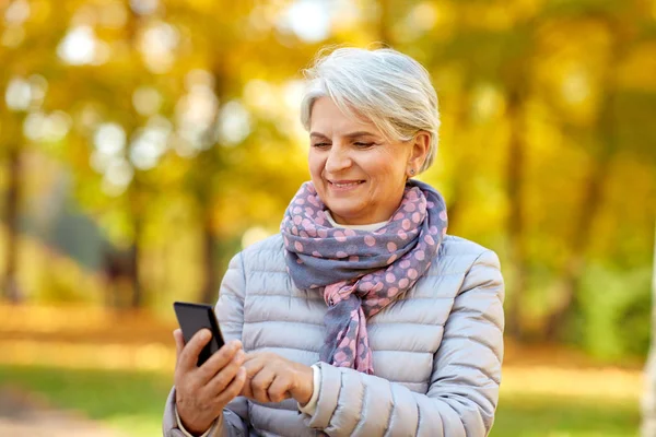 Счастливая пожилая женщина со смартфоном в осеннем парке — стоковое фото