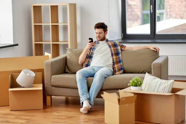 Hombre con smartphone y cajas en el sofá en el nuevo hogar —  Fotos de Stock