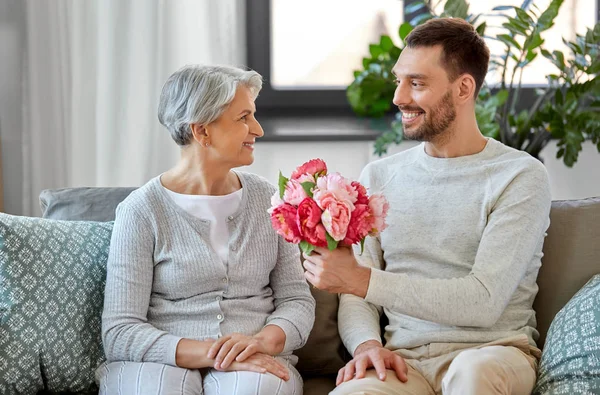 Filho adulto dando flores para a mãe sênior em casa — Fotografia de Stock