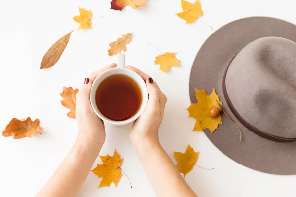 Χέρια με φλιτζάνι τσάι, φθινοπωρινά φύλλα και καπέλο — Φωτογραφία Αρχείου