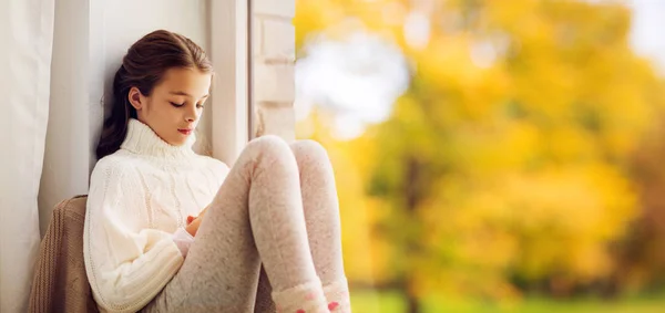 Sad girl sitting on sill at home window in autumn — Stock Photo, Image