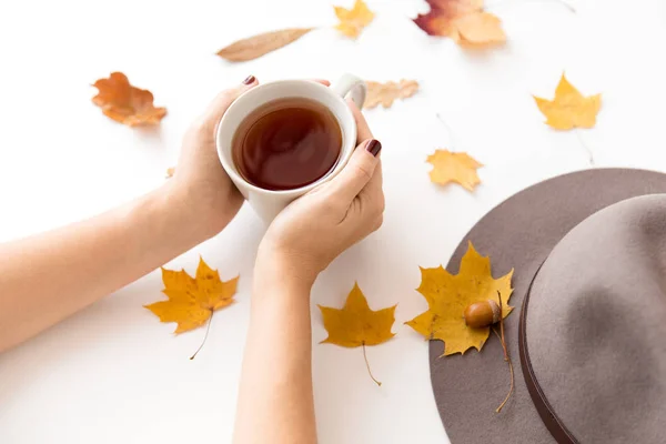 Χέρια με φλιτζάνι τσάι, φθινοπωρινά φύλλα και καπέλο — Φωτογραφία Αρχείου