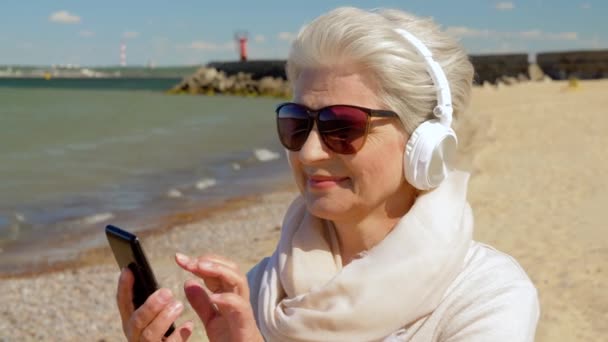 Old woman in headphones with smartphone on beach — Stock Video