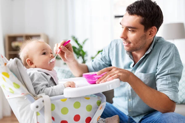 Отец кормит счастливого ребенка в высокий детский стульчик на дому — стоковое фото