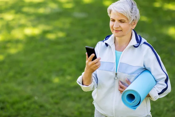 Γριά γυναίκα με χαλάκι άσκησης και smartphone στο πάρκο — Φωτογραφία Αρχείου